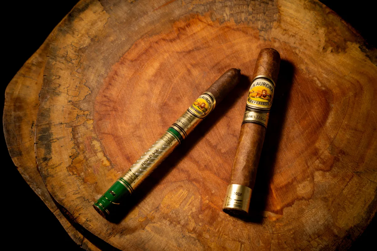 Two cigars, one slender with a green label and one stout with a gold label, resting on a wooden surface.